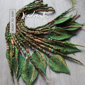 A set of green and brown felted hair dreadlocks featuring attached felt leaves, wooden beads, and metallic accents. The text "diadreads" and "READY TO SHIP!" is visible on the image. The dreadlocks have a textured, twisted appearance.