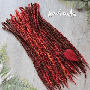 May include: A collection of synthetic dreadlocks in shades of dark brown, rust, and bright orange. The strands feature a twisted texture with small beads and a single red felted leaf accent. The text "diadreads" is visible in white script.