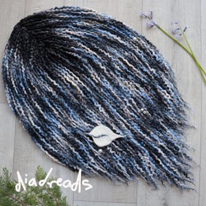 Könnte beinhalten: Nahaufnahme eines Sets blauer, schwarzer und weißer synthetischer Dreadlocks mit silbernen Perlen. Die Dreadlocks sind in einem Kreis auf einer weißen Oberfläche angeordnet. Der Text "diadreeds" ist in der linken unteren Ecke des Bildes sichtbar.