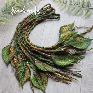 May include: A set of green and brown felted hair extensions featuring leaf-shaped accents, braided textures, and small metallic beads. The text "diadreads" is visible in the upper left corner. The extensions are arranged in a curved shape.