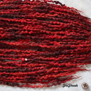 May include: A close-up of a bundle of red and burgundy wool dreadlocks. The dreadlocks are arranged in a wavy pattern and have a natural, textured look.