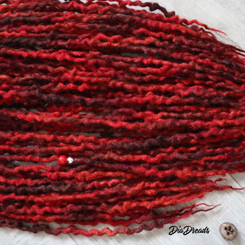 May include: A close-up of a bundle of red and burgundy wool dreadlocks. The dreadlocks are arranged in a wavy pattern and have a natural, textured look.