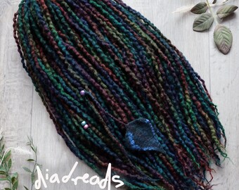 May include: A set of synthetic dreadlocks featuring a blend of dark green, deep purple, and black fibers. The strands are textured and twisted, accented with small beads and a single dark blue leaf-shaped charm. The text "diadreads" is visible.