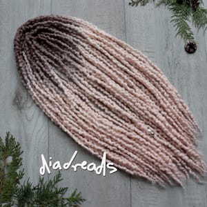 May include: A bundle of handmade dreadlocks in a gradient of brown to light pink. The dreads are arranged on a light gray wooden surface, with the text "diadreads" in white script. A small pearl bead is attached to the dreads.