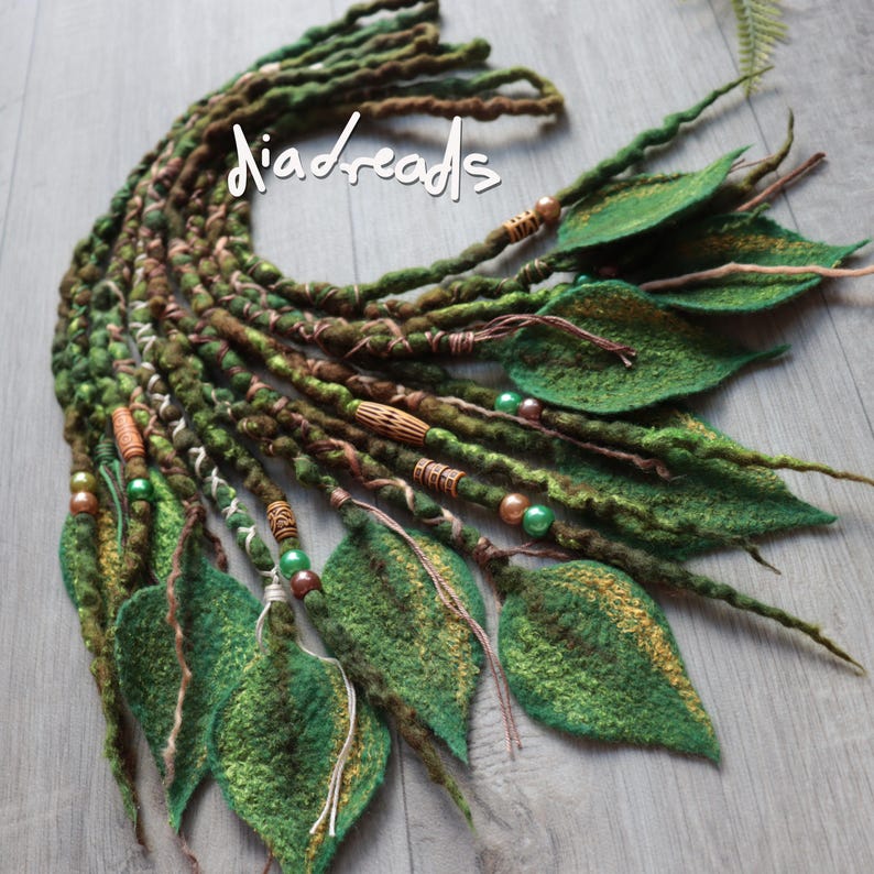May include: A set of green and brown textured dreadlocks featuring the text "diadreads" in white. The dreadlocks are adorned with various wooden and metallic beads, along with several felted leaf-shaped attachments in shades of forest green.