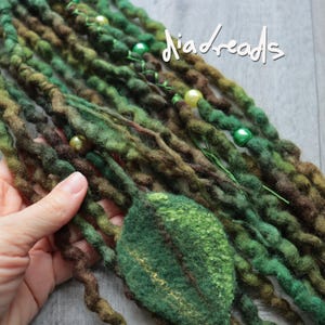 May include: A collection of green and brown felted dreadlocks featuring the text "diadreads" in white script. The set includes textured wool strands adorned with small round beads in shades of lime and olive green, and a large felted leaf accent.