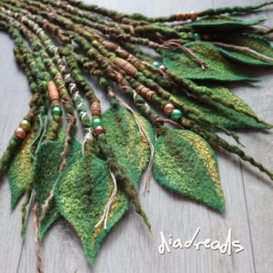 May include: A set of green and brown felted dreadlocks featuring leaf-shaped attachments, wooden and metallic beads, and decorative thread wraps. The text "diadreads" is visible in the bottom right corner of the image.