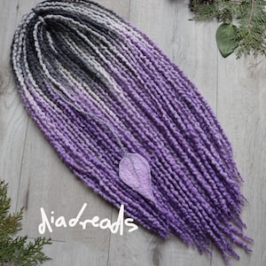 May include: A set of synthetic dreadlocks featuring an ombre color transition from dark charcoal gray at the roots to a vibrant purple at the tips. The textured strands are arranged in a fan shape, with a single felted leaf accent. Text reads diadreads.