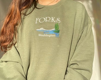 Puede incluir: Una sudadera verde con el texto "FORKS Washington" y una pequeña imagen bordada de un lago y un árbol.