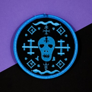 May include: Blue embroidered patch with a skull and crossbones design on a black background. The skull is surrounded by a circle of symbols and a wavy line.