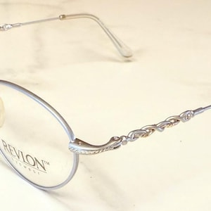 May include: Silver-toned Revlon eyeglasses with oval lenses. The frames feature a delicate, twisted design on the temples and arms. The lenses are clear, and the nose pads are a light yellow color. The Revlon logo is visible on the lens.