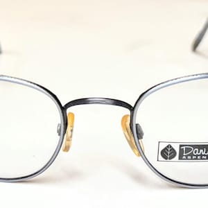 May include: Oval-shaped eyeglasses with a dark gray metal frame. The lenses are clear, and the nose pads are a light yellow color. The brand name "Daniel Hunter Aspen Colorado" is printed on the right lens.