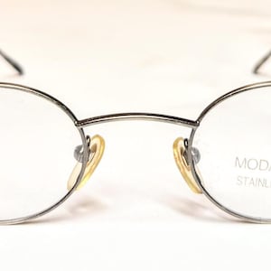 May include: Silver-framed spectacles with oval lenses. The glasses have a thin metal frame, a bridge, and temples. The text "MODA ITALIA STAINLESS STEEL" is printed on the right lens. The nose pads are yellow.
