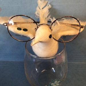 Puede incluir: Un par de gafas redondas con montura dorada y lentes transparentes. Las gafas tienen una bisagra de resorte y el texto "CLIP ON AVAILABLE" y "SPRING HINGE" en la lente izquierda. El texto "CHEROKEE" y "KR-245" está en la lente derecha. Las gafas las lleva puestas una figurita de pájaro blanco y plumoso.