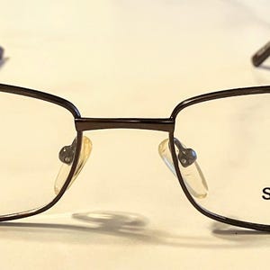 Könnte beinhalten: Rechteckige Brille mit dunkelbraunem Metallrahmen. Die Gläser sind klar, die Nasenpads beige. Auf dem rechten Glas steht "SMART COLLECTION". Die Rahmengröße beträgt 53 mm x 19 mm.