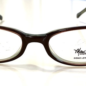 Op de afbeelding: Bruine ovale bril met een donkergrijs montuur. De glazen zijn helder en hebben de tekst "Marc Jacobs" en "DEMO LENS ONLY" in zwart.