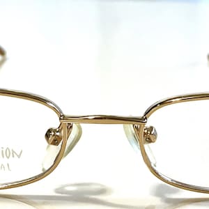 Pode incluir: Óculos de metal dourado com armação retangular e lentes transparentes. Os óculos têm o texto "Eclipse Collection Star Bright Optical" na lente esquerda.