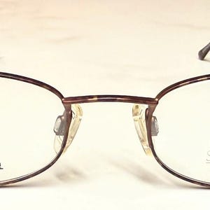May include: Oval spectacles with a brown metal frame and slender, dark temples. The lenses are clear, and the frame is marked "STAINLESS STEEL" on one side and "SYNTHES-EYEZ by Charthant PAT P" on the other.