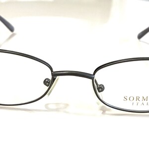 May include: Oval-shaped eyeglasses with a dark gray metal frame. The lenses are clear, and the temples are thin and extend to the ear. The brand name "SORMANI ITALY" is printed on the right lens.