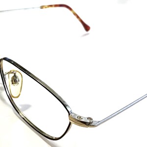 Könnte beinhalten: Silberne und goldfarbene Brille mit einem Schildpatt-Bügel. Die Brille hat einen rechteckigen Rahmen und klare Gläser.