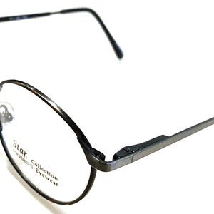 May include: Black metal-framed spectacles with a round lens shape. The glasses have a thin frame and temples, with a Star Collection logo on the lens. The nose pads are a light beige colour.