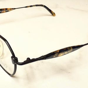 Könnte beinhalten: Schwarze Brille mit geometrischem Rahmen. Die Bügel weisen ein Schildpattmuster mit goldenen Akzenten auf. Die klaren Gläser sind vor einem weißen Hintergrund platziert.