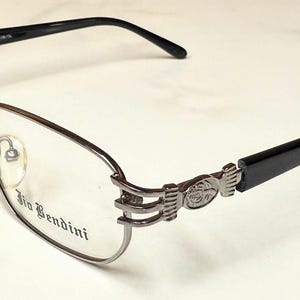 Puede incluir: Gafas con montura plateada y patillas negras. Las gafas tienen forma rectangular con un diseño decorativo en las patillas. La marca "Bio Bendini" está impresa en la lente.