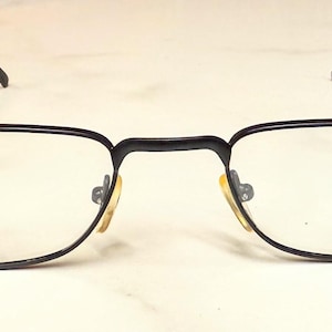 May include: Black rectangular eyeglasses with clear lenses. The frames are thin and made of metal. The nose pads are yellow. The glasses are set against a white background.