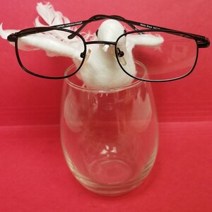 Puede incluir: Una figurita de paloma blanca con gafas de montura negra, sentada en un jarrón de vidrio transparente. Las gafas tienen el texto "Caitlynn Collection Fashion" y "Mary Black 54/17/135" en las patillas.