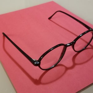 以下が含まれることがあります： ピンクの表面に置かれた、クリアなレンズの黒い丸いフレームの眼鏡