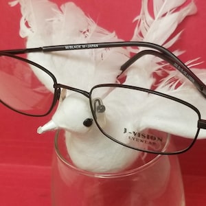 Puede incluir: Gafas rectangulares de marco negro con lentes transparentes. Las gafas están sobre una figurita de pájaro blanco con plumas en un vaso. Las gafas tienen el texto "J-VISION EYEWEAR" y "M/BLACK M-JAPAN" impreso en las monturas.