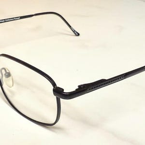 May include: Black metal eyeglasses with rectangular lenses. The frames have a thin, modern design with a textured detail on the temples. The lenses are clear, and the nose pads are visible. The glasses are set against a white background.