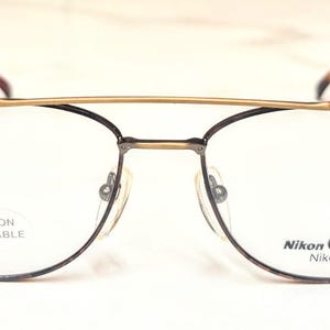 Puede incluir: Gafas Nikon Classic con una barra superior dorada y montura de metal oscuro. Las lentes transparentes muestran el texto "CLIP-ON AVAILABLE" y "Nikon Classic Nikonflex".