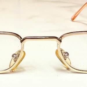 Könnte beinhalten: Goldgerahmte Brille mit rechteckigen Gläsern. Die Fassungen haben ein dünnes, metallisches Design mit Steg und Bügeln. Die Gläser sind klar, und der Text "Scarlet" ist auf einem Glas sichtbar. Die Ohrenstücke sind hellorange.