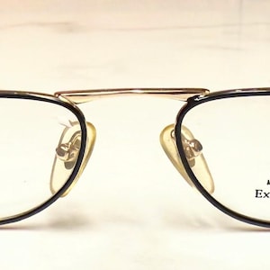 May include: Gold-rimmed eyeglasses with black accents and clear lenses. The frames have a rectangular shape with a double bridge and black temples. The text "Star Collection Exceptional Eyewear" is printed on the lens.