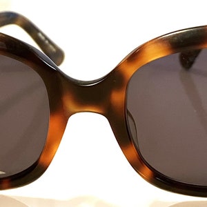 May include: Tortoiseshell sunglasses with dark lenses. The sunglasses have a rounded, oversized frame.