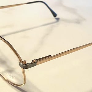 May include: Gold-tone eyeglasses with a rectangular frame and clear lenses. The glasses have a thin, delicate design with black accents on the temples. The frames are made of metal and have a classic, vintage style.