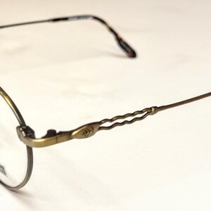 May include: Oval-framed eyeglasses with a bronze-colored metal frame. The temples have a decorative, wavy design and end in a tortoiseshell pattern. The lenses are clear, and the nose pads are beige.