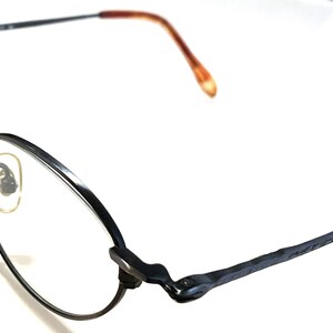 Könnte beinhalten: Schwarze Metallgestellbrille mit runden Gläsern. Die Bügel der Brille haben eine braune Kunststoffspitze.