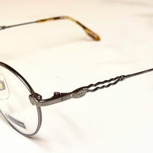 May include: Oval-shaped eyeglasses with a dark metal frame. The temples have a decorative twist design and a leaf accent. The earpieces are a tortoiseshell pattern. The lenses are clear.