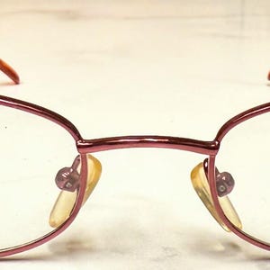 May include: Oval-shaped eyeglasses with a burgundy frame and temples. The lenses are clear, and the nose pads are yellow. The text "CLUB MAD" is printed on the right lens. The glasses are designed for vision correction.