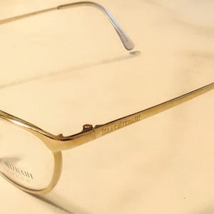 Op de afbeelding: Goudkleurige bril met een dun montuur en heldere glazen. De merknaam "ISAAC MIZRAHI" is zichtbaar op de poot. De bril heeft een klassiek, elegant ontwerp.