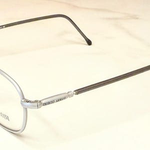 May include: Silver-framed eyeglasses with a thin, dark gray metal arm and a clear lens. The brand name "Giorgio Armani" is visible on the arm. The glasses have a classic, minimalist design.