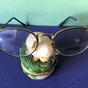 Può includere: Occhiali ovali con montatura dorata e aste verdi, appoggiati su una piccola statuetta di ceramica di una pecora bianca. Gli occhiali hanno la scritta "Fundamentals" stampata sulla lente destra.