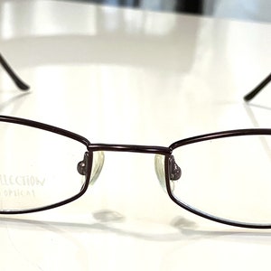 Könnte beinhalten: Eine Brille aus braunem Metall mit einem rechteckigen Gestell und klaren Gläsern. Die Brille hat den Text "allure collection the bridge optical" auf dem rechten Glas.