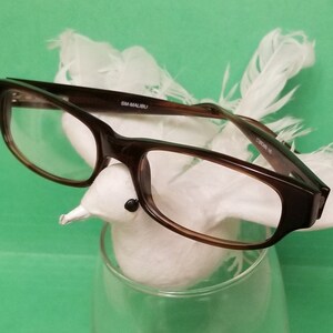 May include: Brown eyeglasses with a white feathered bird perched on the frame. The bird is sitting in a clear glass.