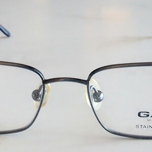 May include: Rectangular eyeglasses with a dark gray metal frame. The lenses are clear, and the brand name "GANT" is printed on the right lens. The frame is made of stainless steel.