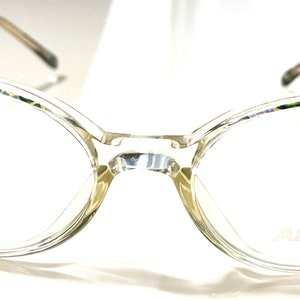 Peut inclure: Lunettes à monture transparente de forme œil de chat. Les montures présentent un motif irisé subtil sur le dessus et les côtés, avec des accents dorés décoratifs aux tempes. La marque "ARISTAR" est visible sur le verre.