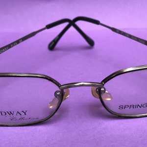 Può includere: Occhiali in metallo color argento con cerniera a molla. Gli occhiali hanno una forma rettangolare e riportano la scritta "BROADWAY Collection" sulla lente sinistra e "SPRING HINGE" sulla lente destra.