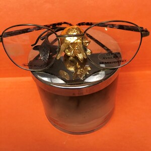 Puede incluir: Un objeto decorativo de metal dorado con un diseño de pájaro se coloca encima de un frasco de vidrio con una tapa plateada. El frasco contiene una sustancia marrón. Un par de gafas de pasta de carey marrón con bisagras de resorte y el texto "Clip on Available Members Only" descansan sobre el frasco.